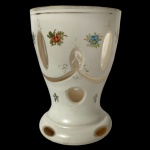 Copo em grosso cristal da Boêmia, de formato cônico, revestido por overlay leitoso. Borda decorada por filete dourado. Apresenta lapidação com pequenas janelas sobre guirlanda com arcos, realçada por adornos florais policromados. Base circular, saliente e esparramada com borda ornada por facetas ovais. 11.5 cm.