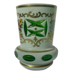 Copo em grosso cristal da Boêmia de cor verde esmeralda, revestido por overlay leitoso e frisos a ouro. Bojo cônico e reto, lapidado e ornado por janelas geométricas, alternadas com arranjos florais policromados e bordadas por volutas douradas. Base anelada e saliente, realçada por adornos ovais, apoiada em fundo retraído. 9 cm.