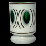 Copo em grosso cristal da Boêmia de cor verde esmeralda, revestido por overlay leitoso e adornos a ouro. Bojo cilíndrico reto, lapidado e ornado com janelas ovais, alternadas por traços recurvos. Base circular almofadada lisa. 6.5 cm.