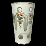 Copo em grosso cristal transparente da Boêmia, de formato cônico reto, revestido por overlay leitoso e realçado por bordas recortadas em pequenos arcos. Bojo lapidado e ornado com facetas ovaladas, alternadas por arranjos florais policromados. Base com pequenas janelas circulares. 13 cm.