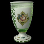 Copo em grosso cristal da Boêmia de cor verde esmeralda, revestido por overlay leitoso e frisos a ouro. Bojo cônico, facetado e ornado com treliças verticais alternadas por arranjos florais policromados. Apresenta bordas lapidadas com anelados. Base apoiada em pé circular, acompanhando o estilo com friso inferior dentado. 13.5 cm.