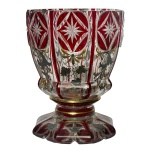 Copo em grosso cristal transparente da Boêmia, com laterais levemente arqueadas, revestido e ornado por overlay de cor rubi. Apresenta rica ornamentação com painéis verticais, decorados por adornos vegetais encimados por estrelas lapidadas e emolduradas. Base com anelado saliente, apoiada em aros retraídos e dourados. Pé circular esparramado e recortado em pétalas. Séc. XIX. 5 cm.
