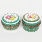 PORCELANA, duas (2) caixas miniatura: 1 ornada com cena de galanteio; 1 ornada com flores, ambas em policromia com arremates a ouro e monture em bronze, fecho de uma delas partido, marca de manufatura na base, medindo em média 6 cm diâmetro e 4 cm altura, vide foto.