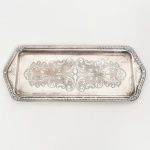 AÇO INOX, uma (1) travessa quase retangular, fundo em baixo relevo representando volutas e elementos vegetalistas, borda relevada em corda, conchas e elementos vegetalistas, sem marca de manufatura aparente, medindo 33 x 14 cm sinais de uso, vide foto.