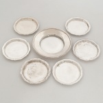 AÇO INOX E METAL PRATEADO, sete (7) itens: 1 porta garrafa, sem marca de manufatura aparente, medindo 12 cm diâmetro; 6 porta copos com borda plissada e recortada, manufatura CRISTOFOLI, medindo 8,5 cm diâmetro, sinais de uso, vide foto.