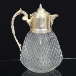 VIDRO, uma (1) jarra com modelagem geometrizada, incolor e translúcida, formato quase piramidal, monture e alça em metal prateado, relevada em perolados e elementos vegetalistas, interior com recipiente separador de gelo, selo da Roberto Simões, medindo 26 cm altura, sinais de uso, vide foto.