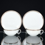 PORCELANA, par (2) xícaras para chá, tonalidade branca, arremate com friso prateado, modelo MEDAILLON, manufatura RENNER (Porto Alegre, RS, fundada em 1942; em 1998 foi vendida para a Vista Alegre de Portugal; em 1999 mudou a razão social para Vista Alegre do Brasil), ambas as xícaras com bicados na borda, uma delas com trincado acentuado, sinais de uso, vide foto.