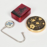 DIVERSOS, três (3) itens: 1 trousse confeccionada em metal dourado, tampa ornada com pássaro e flores na técnica CHOKIN (gravação em cobre, prata e ouro, tradição que vem do século XII, período dos Samurais), interior com espelho, medindo 7,5 cm diâmetro; 1 suporte para pendurar objetos com propaganda do Cursos CNF, medindo 9 cm, confeccionado em metal e outros materiais; 1 caixa em laca vermelha e preta, ornada com silhueta figurativa, medindo 7 x 5 x 3 cm, trincada na base, todos os itens com sinais de uso, vide foto.