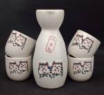 Conjunto de saquê, em porcelana, sendo uma garrafa e quatro copos com figura estampada de gato da sorte. Medindo a garrafa com 12 cm e copo com 4 cm.