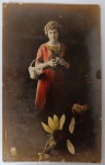 Cartão-Postal Antigo Colorizado  Retrato de Moça com Cesto de Flores (Cerca de 1910)Imagem de uma jovem mulher em traje de época, segurando um cesto com flores e uma margarida nas mãos. Em primeiro plano, destaca-se a sobreposição artística de uma grande flor amarela.