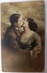 Antigo Cartão Postal "O Beijo"  Fotografia de Estúdio Colorizada à Mão (Circa 1910-1920)Raro cartão postal antigo apresentando uma cena romântica de um casal em um abraço afetuoso. Este tipo de postal era extremamente popular durante a Belle Époque e o período da Primeira Guerra Mundial, frequentemente trocado entre apaixonados