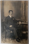 Fotografia Original Antiga (Cartão-Postal)  Retrato Masculino de Época  Datada 1911Retrato de estúdio de um jovem cavalheiro trajando terno de gala, colarinho alto e bigode, característicos da moda masculina de 1910. Composição clássica com mobiliário de época e fundo cenográfico pintado