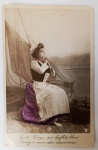 Antigo Postal Centenário (1900-1915)  Fotografia Colorizada "Protegei meu amado"Cena romântica e dramática de uma jovem mulher em trajes de camponesa/pescadora, sentada à beira-mar com um barco a vela ao fundo. Ela está em posição de oração, olhando para o céu.