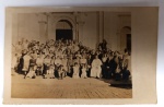 Raro Postal Fotográfico  Fiéis na Basílica Velha de Aparecida (SP)  Circa 1950Registro de um grande grupo de devotos e um sacerdote (provavelmente Redentorista, ordem que administra o santuário desde 1894) posando em frente ao portal da Basílica Histórica. É um documento valioso sobre as romarias e a cultura material religiosa do meados do século XX.