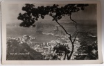 Antigo Postal Fotográfico Rio de Janeiro  Vista Panorâmica Enseada de Botafogo e Pão de Açúcar  Foto Lucarelli  Circa 1950Composição artística emoldurada por galhos de árvores em primeiro plano, revelando a Enseada de Botafogo, a Urca e o Morro do Pão de Açúcar ao fundo. Um registro icônico da topografia carioca antes da intensa verticalização da zona sul.Identificação: Legenda manuscrita na chapa: "RIO DE JANEIRO" (esquerda) e "FOTO LUCARELLI" (direita)