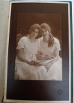 Retrato de Duas Jovens com Livro - Ateliê Frenzel & Schmidt - São Paulo (c. 1915-1920) Fotografia original de época em suporte de cartão rígido texturizado. Apresenta duas jovens em trajes de gala brancos, com ricos detalhes em renda e cintos ornamentados, segurando um livro. A iluminação de estúdio ressalta os penteados típicos do período e a expressão serena das retratadas.