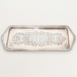 METAL PRATEADO, uma (1) bandeja para rocambole, fundo com ornamentação em baixo relevo representando volutas e elementos vegetalistas, borda relevada em corda, conchas e elementos vegetalistas, medindo 33 x 14 cm, sinais de uso, vide foto.