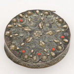 METAL PRATEADO, uma (1) antiga caixinha com tampa relevada em elementos vegetalistas, apliques em cabochões de coral ou símile, faltando alguns, medindo 7 cm diâmetro, sem punções aparentes, sinais de uso, mossas, vide foto.