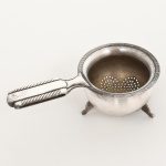 METAL PRATEADO, um (1) suporte individual para infusão de chá, borda serrilhada, base trípode, sem marca de manufatura aparente, medindo 11 cm comprimento, sinais de uso, vide foto.
