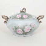 PORCELANA, uma (1) terrina oval com tampa, ornamentação pintada a mão pelo Atelier IOLANDA 1971, representando rosas em policromia sobre fundo na tonalidade azul, alças e pega da tampa com arremates prateados, marca SCHMIDT (Pomerode, SC; Mauá, SP; Campo Largo, PR, fundada em 1945, ainda em atividade, maior fábrica de porcelana da América Latina), medindo 31,5 x 19 x 19 cm, vide foto