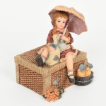 DIVERSOS, uma (1) caixa com tampa, confeccionada em resina policromada, representando menina com patos e sombrinha, além de regador, buque de flores e bacia, medindo 13 cm altura, vide foto.