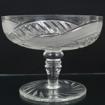 CRISTAL, uma (1) fruteira de pé alto, incolor e translúcida, lapidada em folhas e sulcos, formando padrões geometrizados, fuste no formato de coluneta, base em bolacha, medindo 25 cm diâmetro e 19 cm altura, sinais de uso, vide foto.