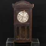 RELÓGIO, um (1) de parede, marca JUNGHANS (Gebr. Junghans Uhrenfabrik, fundada em 1861, por Erhard Junghans, inicialmente como fabricante de peças para relógios; em 1866 produziu seu primeiro relógio por completo; em 1870 morre Erhard Junghans e seus filhos Arthur e Erhard Jr assumem a fabrica; em 1877 fez o registro da primeira marca da fabrica representada por uma águia com uma bandeira; em 1890 registrou a marca com a estrela de oito pontas; em 1903 já era o maior fabricante de relógios do mundo; depois da Guerra o bisneto Helmut Junghans dá continuidade aos negócios da família; em 1927 começou a produzir relógios de pulso; em 1956 passou a ser propriedade da Diehl; em 1985 lança o primeiro relógio atômico; em 1986 lança o primeiro relógio solar atômico; em 1989 lança o primeiro relógio de pulso atômico; em 1991 lança o primeiro relógio atômico com movimento analógico com antena externa; em 1994 idem com antena integrada), mostrador em metal com algarismos arábicos, com dois furos para dar corda, falta o pendulo e a chave borboleta, gabinete em madeira e placagem com porta envidraçada, placagem com falhas, entalhe vegetalista, medindo 69 cm altura, usado, não testado e sem garantia, vide foto.