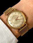 JAEGER-LECOULTRE EM OURO 18K - Relógio masculino Jaeger-LeCoultre, modelo Memovox, exemplar de notável elegância e refinamento técnico, produzido circa fins da década de 1950 / início dos anos 1960. Caixa em ouro amarelo 18 quilates (750), medindo 35 mm, acompanhada de pulseira tipo malha igualmente em ouro, compondo conjunto de presença sólida e sofisticada ao pulso. Mostrador prateado com centro rebaixado e disco interno rotativo de alarme, solução característica da linha Memovox, aqui indicado por discreto triângulo azul, elemento funcional que se transforma em assinatura estética. Índices aplicados e ponteiros tipo dauphine reforçam a linguagem clássica do período.Equipado com movimento mecânico de corda manual dotado de função despertador  complicação que consagrou a manufatura e a colocou entre as grandes referências da relojoaria do século XX. *ITEM PARA COLECIONADOR.Verso gravado Memovox, confirmando a natureza do modelo.Peça que conjuga engenharia e poesia mecânica, representando uma era em que o relógio de pulso transcendia a medição do tempo para dialogar com o cotidiano de seu portador despertando-o, literalmente, para o mundo.