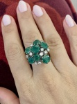 ANEL EM PLATINA COM ESMERALDAS E DIAMANTES. Anel em platina, de desenho volumoso e composição floral, centrado por sete esmeraldas naturais de lapidação oval mista, totalizando aproximadamente 4,0 ct, dispostas em cluster elevado, formando conjunto de forte presença visual. Entre as gemas verdes, distribuem-se dez diamantes antigos (lapidação tipo 8/8), com peso estimado total de aprox. 1,4 ct, conferindo contraste luminoso e leitura clássica à peça. Montagem com garras altas e estrutura vazada inferior característica de joalheria de meados do século XX privilegiando a incidência de luz e o volume escultórico. As esmeraldas apresentam coloração verde média a intensa, com inclusões naturais visíveis (jardim), compatíveis com a origem da gema e sem prejuízo ao conjunto estético. Aro: 18. Peso: 9,45 g. Material: platina. Pedras: 7 esmeraldas ( 4,0 ct total) e 10 diamantes ( 1,4 ct total).