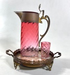 ANTIGA E PEQUENA JARRA EM VIDRO VERMELHO  COM GUARNIÇÃO EM METAL E COPINHO, BANDEJA REDONDA EM METAL COM BASE DE ESPELHO COM DESGASTE. MEDIDAS: JARRA 17 CM / COPINHO 5 CM / BANDEJA 13,5 CM DE DIÂMETRO.