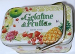 Lata Tipo Maleta "Gelatine di Frutta"  Itália (C. 1960/70)Encantadora lata litografada vintage para Gelatine di Frutta (gelatinas de frutas), com o raro formato de "maleta" ou marmita, incluindo alça metálica móvel e tampa de encaixe profundo. O design apresenta uma composição colorida e detalhada de frutas (maçã, abacaxi, morangos e cerejas) sobre um fundo creme com padrões florais em tons pastéis.