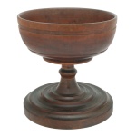 Centro de mesa em madeira, lustrada. Apresenta formato de pedestal com uma cuba superior larga apoiada sobre uma coluna torneada e base circular em degraus. 27 x 27 cm. Na tradição europeia, tigelas desse formato eram usadas para servir bebidas festivas quentes em celebrações (como o wassail no inverno).