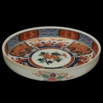 Saladeira circular com fundo plano em porcelana japonesa, decorado em estilo Imari com esmaltes rouge de fer, azul cobalto, verde e ouro. Apresenta parte interna com borda realçada por reservas estilizadas, centralizando margem com painéis floridos e rosácea ao meio. Parte externa com discretos adornos. Marca do ceramista sob a base. 6 x 28 cm.