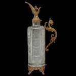 Belíssimo licoreiro de formato cilíndrico em cristal lapidado. Apresenta guarnições em bronze dourado e alça em forma de dragão. 40 x 16 cm.