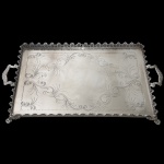 Grande bandeja em metal prateado. Formato retangular com cinzelados, centro com decoração de arabescos e folhagens e borda com concheados. Pés de garra e alças trabalhadas. 7 x 70 x 40 cm.