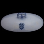 Tampa de sopeira de formato oval em porcelana brilhante em tons de azul. Apresenta um monograma intrincado em azul no centro da tampa, A pega é estilizada com formas geométricas e detalhes lineares em azul. 8 x 25 x 16 cm.