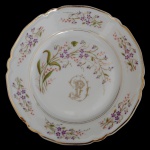 Prato em porcelana provavelmente francesa. decorado com ramos de flores lilás e folhagens verdes espalhadas pela borda e centro. Borda ondulada é realçada com um acabamento em ouro. Centro exibe um monograma dourado estilizado. 21 cm.