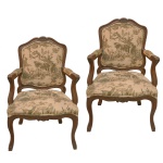 Par de cadeiras de braços estilo Luís XV (frequentemente chamada de fauteuil). O design destaca-se pelas linhas curvas fluidas, pernas estilo cabriole (pés de cabra) e entalhes ornamentais na madeira decorando o topo do encosto e a saia inferior. O estofado apresenta uma estampa clássica em estilo Toile de Jouy, caracterizada por cenas pastoris e campestres monocromáticas em tons de verde sobre um fundo claro.. 97 x 64 x 52 cm.