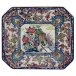 Travessa retangular em porcelana japonesa estilo Imari com cantos chanfrados e decoração de carroça de flores. Apresenta um padrão pictórico colorido de uma carroça de flores (rickshaw) no centro, rodeada por motivos florais e figuras em tons de azul, vermelho e verde. 30 x 33 cm.