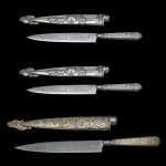 Conjunto de três facas gaúchas, com bainha com detalhes artesanais específicos. Apresentam bainhas de metal prateado com relevos que retratam cenas rurais, incluindo carroças e figuras a cavalo. Uma das facas exibe bainhas e cabos decorados com motivos florais detalhados. 24 cm e 28 cm.