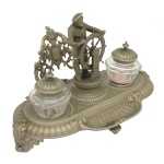 Tinteiro de mesa em metal prateado repuxado e cinzelado, com bandeja oval moldada por bordas em gomos, interrompida à frente por descanso escavado para canetas. Apresenta figura de timoneiro com as mãos no leme, encostado em adorno estilizado e vazado com escudo ao centro, e nas laterais, recipientes em vidro com tampas. 20 x 30 x 18 cm.