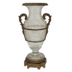 Vaso de formato balaústre em cristal transparente, lapidado e ornado por faixas geométricas centralizando larga barra em bico de jaca. Gargalo facetado em corneta e base circular. Montagem em bronze com bordas e frisos laterais sustentando alças rematadas por volutas, e peanha quadrangular com quinas recortadas. Séc. XIX. 57 x 30 cm.