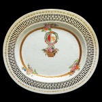 Travessa ovalada em porcelana chinesa da Companhia das Índias, decorada em esmaltes policromados. Apresenta fina borda estilizada e larga margem vazada. Fundo ornado por faixa florida, e cesta com flores sob duplo brasão de armas e coroa. Início do séc. XIX. Necessita de restauro na borda.