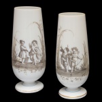 Par de vasos floreiros de formato alongado, em opalina leitosa, ornamentadas em sépia por cenas de pescaria com crianças e anjos. Bojos no formato de copos, apoiados em bases circulares lisas e ornadas por filetes azuis e frisos dourados. 26 x 10 cm.