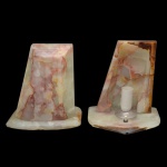 Par de luminárias de mesa, montadas com placas de ônix rajado em tons mesclados. Bases retangulares, com frentes arqueadas e lados posteriores encimados por segmentos recortados em trapézio e escavados. Apresentam ao centro pequenas canoplas rematadas por bocais para lâmpadas. 20 x 17 x 13 cm. Fiação nova.