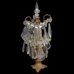 Elegante girandole em bronze ormolu, ornamentada por placas em cristal facetado. Coluna reta e anelada, sustentando braços recurvos distribuídos em 2 níveis, e realçados por pequenas flores lapidadas. Parte superior com pequena urna encimada por ponteira apoiada em argola. Base esparramada e ornada por sulcos lisos rematados por pés lobulados. 50 x 20 cm. Marcas de uso.