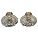 Par de pequenos castiçais baixos em metal prateado repuxado e cinzelado. Pratos circulares com folhagens na borda, centralizando os porta velas. 3 x 7 cm. Peso 74 gramas. porta cigarros de metal na forma de taça. (9 x 7 cm).