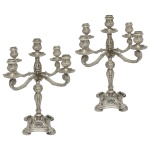 Par de candelabros para 5 velas em prata brasileira repuxada e cinzelada, estilo D. João V, ornados por folhagens, volutas e reservas. Hastes retas abauladas ao centro, encimadas por esferas lavradas. Braços recurvos, sustentando bobeches e centralizando ponteiras com pequenos castiçais no estilo. Bases quadrangulares recortadas com pés esparramados. Contrastes da prata de teor 833 mls e marcas de prateiro de São Paulo. 38 x 31 cm. Peso aprox. 2K235 gramas.
