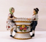 CAPODIMONTE-  Uma floreira em porcelana italiana, representando " Jovens Camponeses e Menino com Menina" em rica policromia, Séc XIX.. Manufatura no verso. Med. 15 x 15 x 9 cm. Perda do dedo indicador nas mãos direita e esquerda , que não interfere na beleza da peça.