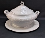 VERBANO - ARGENTINA-Belíssima sopeira com presentoir anos 80, em porcelana branca moldada na pasta com florais. Possui duas alças laterais em guirlandas. Acompanha grande presentoir decorado com florais e frutas. Med. sopeira 25 cm alt x 33 cm diâmetro, presentoir 33 cm diâmetro.